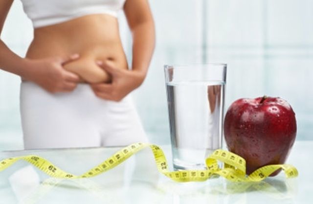 Mujer agarrandose la grasa del abdomen tras una mesa con cintra métrica amarilla,vaso de agua y una manzana