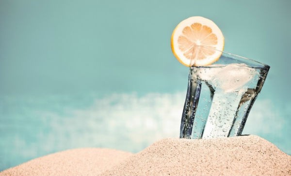 Vaso de cirtsal con agua y limón en arena de la playa con el mar de fondo