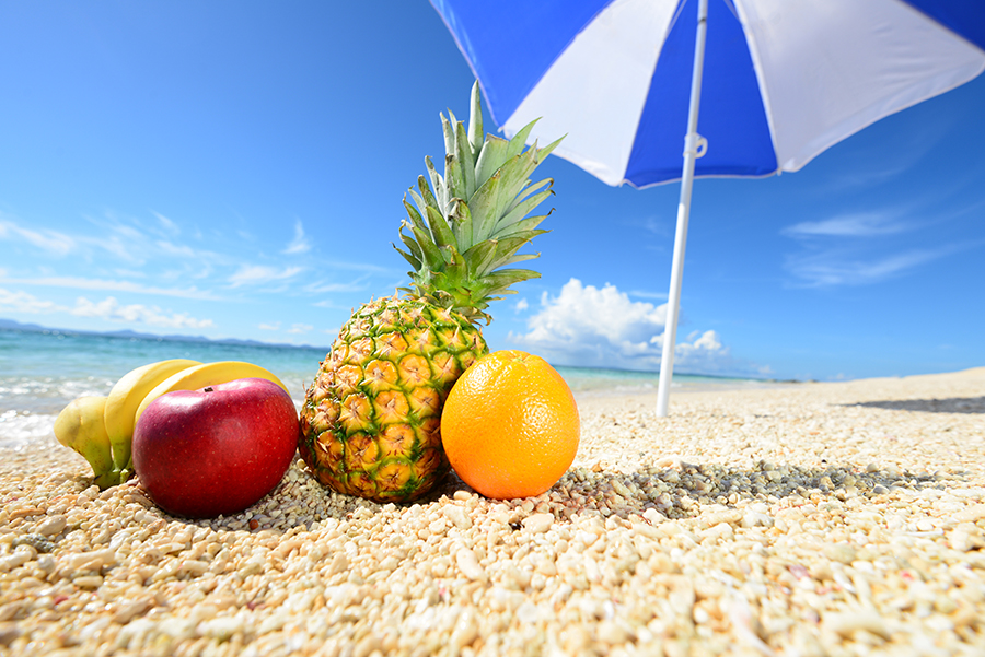 Sombrilla azul y blanca en la playa con frutas como naranja y piña en la arena
